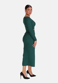 Grünes geripptes Zweiset bestehend aus einem cropped Oberteil mit langen Ärmeln und einem taillierten Midirock. Getragen mit schwarzen Stiletto-Absätzen.