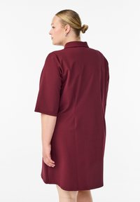 Vrouw met blond haar in een knot draagt een knielange, kortmouwen bordeauxrode jurk en staat zijwaarts tegen een plain witte achtergrond.