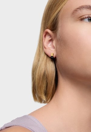 Gros plan sur l'oreille d'une femme aux cheveux blonds courts, portant une boucle d'oreille en forme d'ourson en or avec une petite perle noire en dessous.