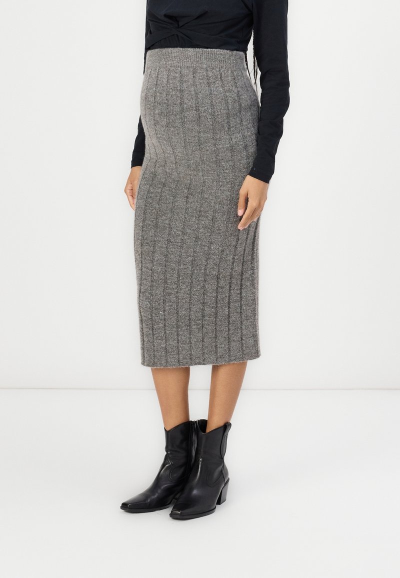 Femme portant une jupe midi en maille côtelée grise taille haute, des bottines noires et un haut noir à manches longues, debout devant un fond blanc.