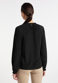 Blouse noire à manches longues avec un col froncé et des poignets élastiques. Texture de tissu lisse, silhouette ajustée et discrète marque à l'arrière.