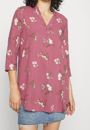 Frau trägt eine mauvefarbene Tunika mit rosa-weißen Blumen- und Schmetterlingsdruck, dreiviertellangen Ärmeln und einen blauen Jeansrock mit ausgefranstem Saum.