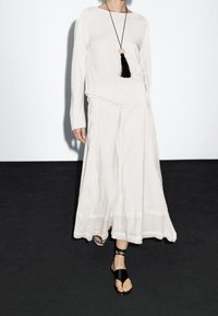 Kermanvärinen pitkähihaiset paita, jossa on suora pääntie, yhdistetty ilmavaan nilkkapituiseen hameeseen. Varustettu mustilla remmisandaaleilla ja riipuskaulakorulla.