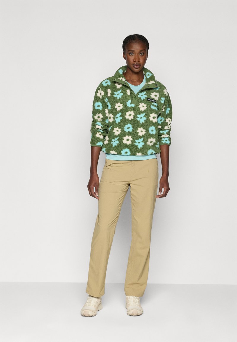 Pullover in pile verde con motivo floreale in bianco e blu, dotato di un colletto alto e zip, abbinato a pantaloni beige e scarpe chiare.