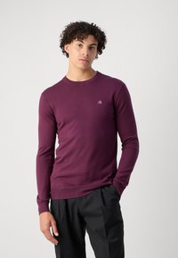 Maglione in maglia borgogna con maniche lunghe, scollatura rotonda e un piccolo logo sul petto. Il materiale sembra morbido con una texture liscia.