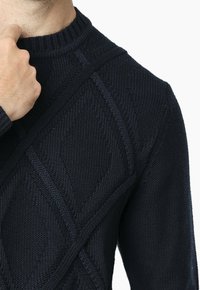 Jersey de marina en punto texturizado, con un patrón de diamantes y un cuello redondo acanalado. Primer plano de la parte superior del torso y una mano cerca del cuello.