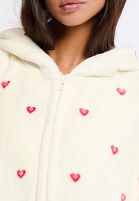 Felpa con zip di colore crema, realizzata in morbido tessuto, caratterizzata da motivi di cuori rossi sparsi, e con cappuccio a collo rotondo.