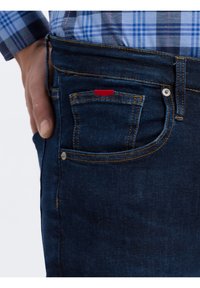 Ruka v zadní kapse tmavě modrých džín s modrou kostkovanou košilí, která ukazuje detailní prošití a kovové nýty.