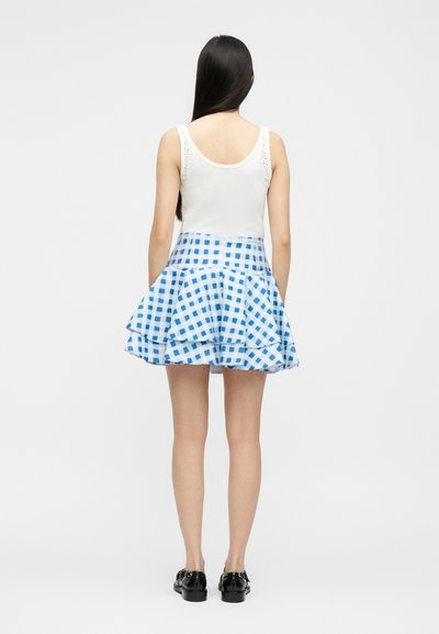 Γυναίκα με μακριά σκούρα μαλλιά που φοράει λευκό αμάνικο τοπ και μπλε-λευκή καρό πολυεπίπεδη φούστα, στεκόμενη με την πλάτη σε λευκό φόντο.