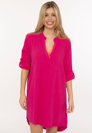 Lächelnde Frau trägt ein knielanges, leuchtend pinkes Kleid mit hochgekrempelten Ärmeln und oranger Einfassung am Ausschnitt, steht vor weißem Hintergrund.