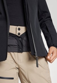 Schwarze Jacke mit Reißverschluss, ausgestattet mit einem beigen Bund, der zwei schwarze Knöpfe und einen strukturierten Stoff aufweist. Beigefarbene Hose hat eine Taschenakzent.