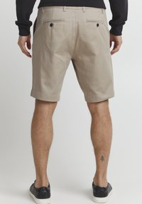 Beige shorts med en tekstureret overflade, som har to baglommer, metalknapper og en lige kant, båret med sorte sneakers.