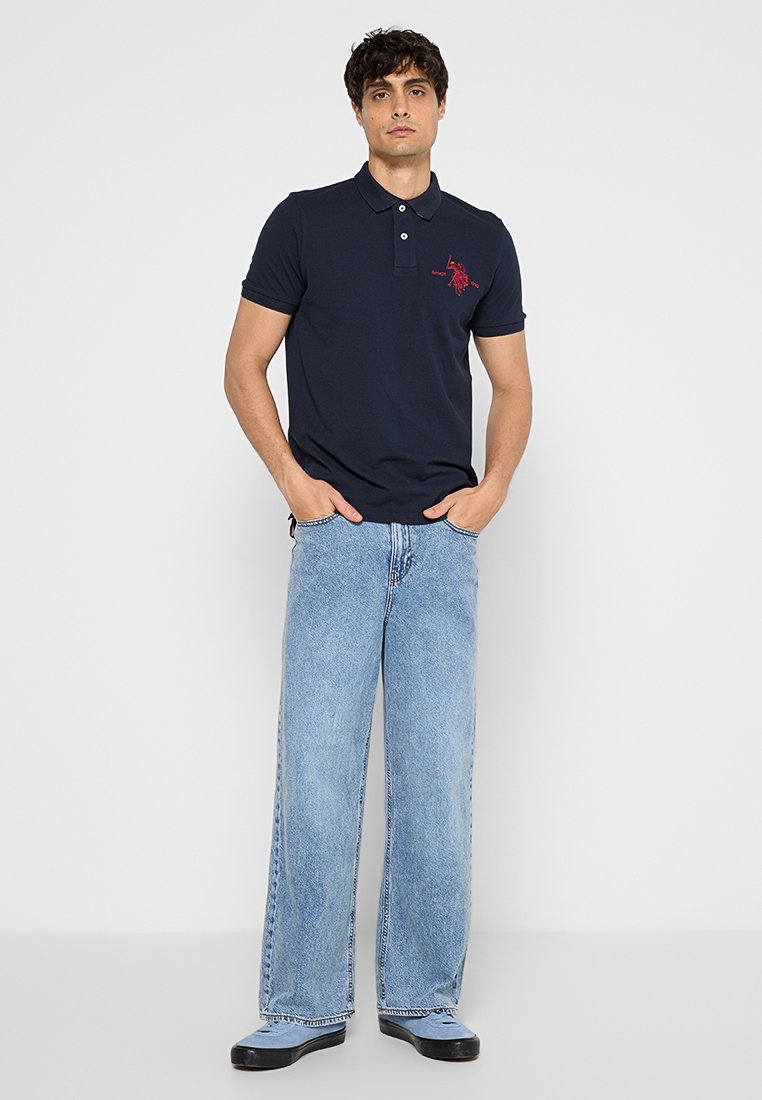 Homme debout, les mains dans les poches, portant un polo bleu marine avec un logo rouge, un jean large bleu clair et des chaussures noires, sur fond blanc.