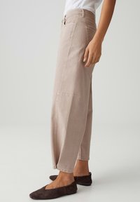 Persona che indossa pantaloni beige a vita alta e gamba larga e scarpe basse marrone scuro, in piedi di lato contro uno sfondo neutro.