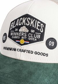 Gorra blanca y verde con parche negro bordado que dice "BLACKSKIES Winners Club Earned Not Given" y "Premium Crafted Goods."