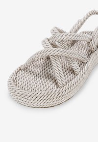 Sandale en corde tressée de couleur beige clair avec un design à brides croisées. Elle présente une semelle intérieure texturée et une semelle arrondie, mettant en avant un esthétique minimaliste.