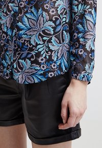 Svart genomskinlig skjorta med blå blommönster och bladmönster, med knäppning fram och uppvikta ärmar, kombinerad med svarta lädershorts.