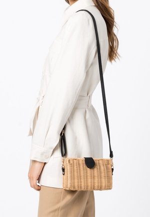 Mujer con chaqueta blanca y pantalones beige, llevando un bolso bandolera rectangular de tejido con correa y solapa negra.