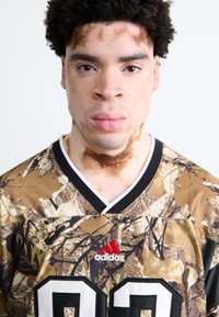 Kamuflage trøje i brune og grønne nuancer med sorte accenter, der har en hvid krave, rødt Adidas-logo og stort sort trøjenummer 02.