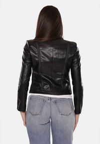 Giacca di pelle nera con design aderente, cuciture strutturate e texture liscia. Abbinata a jeans blu e una camicia bianca.