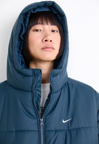 Temnočloveš puffer jakna z visokim ovratnikom in prevelikim kapuco. Ima sprednji zadrgo in bel logotip Nike na levi prsi. Gladka tekstura.