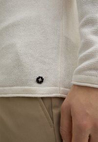 Naturweißer Strickpullover mit einem strukturierten Muster, versehen mit einem schwarzen Logo-Akzent am unteren Saum und einem gerippten Bündchen am Handgelenk.
