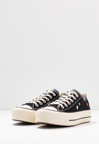 Czarne trampki na płaskiej podeszwie z haftowanymi kwiatowymi detalami, kremową gumową podeszwą i beżowymi sznurówkami. Charakteryzują się klasycznym, niskim krojem.