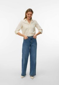 Femme souriante, les mains dans les poches, portant une chemise crème à boutons avec manches retroussées, un jean large bleu et des chaussures blanches à bout pointu.