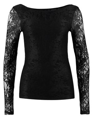 Frau trägt ein schwarzes, langärmliges Spitzenoberteil, verwaschene blaue Jeans mit Umschlägen, schwarze Stiefeletten und hält eine schwarze Clutch-Tasche.