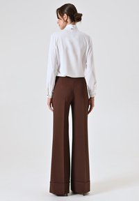 Blouse blanche à manches longues avec col mandarin et poignets boutonnés, portée avec un pantalon large marron à ourlet plié.