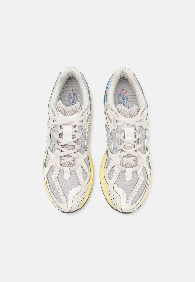 Zapatillas de running gris claro con parte superior de malla, cordones blancos y suela amarilla. Presenta acentos azules en los costados y un diseño estructurado.