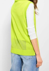 Gilet senza maniche verde lime con un design in rete testurizzata, bordi a coste e un sottile dettaglio sulla tasca, abbinato a una camicia bianca a maniche lunghe.