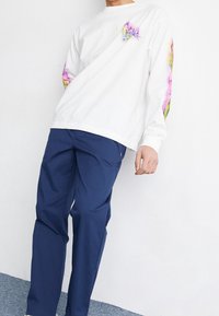 Camicia bianca a maniche lunghe con accenti grafici colorati sulle maniche, abbinata a pantaloni blu scuro. Il tessuto appare liscio e informale.