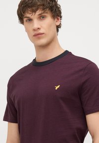 Burgundi lühikese varrukaga t-särk musta ümariku kaelusega, mille vasakul pool on väike kollane linnu embleem. Pehme puuvillane tekstuur.