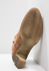 Bruine suède enkellaars met een chunky hak, voorzien van een ronde neus en metallic accenten op de zijknop. De zool heeft een structuurpatroon.