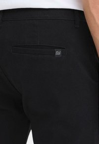 Zwarte broek van geweven stof met een gladde textuur, voorzien van een achterzak en een klein logo tag op de zak.