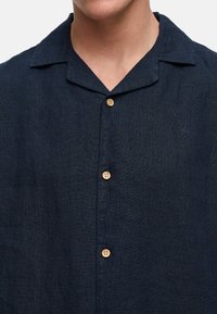 Camisa de lino azul marino con cuello muesca, cinco botones de madera y bordado sutil. La tela tiene un acabado texturizado y un corte relajado.