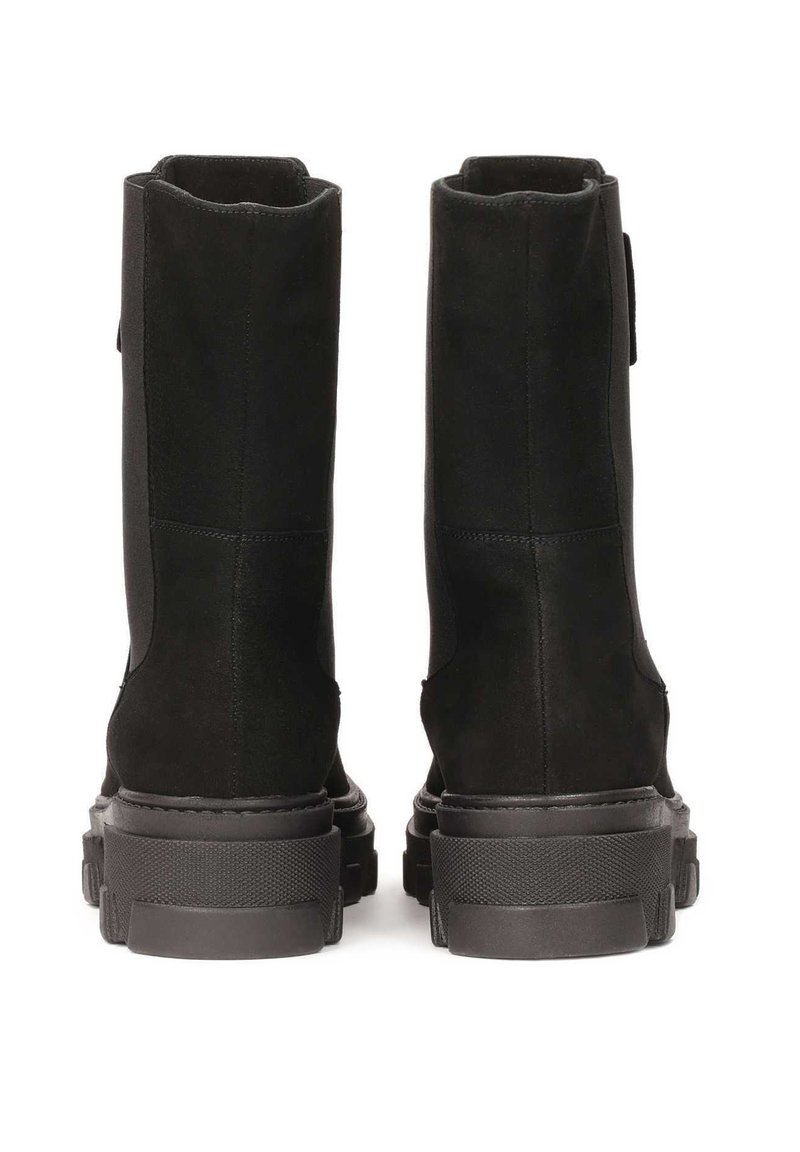 Para czarnych zamszowych botków z masywnymi gumowymi podeszwami, widziana z tyłu na białym tle.