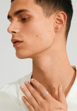 Silver wave-shaped ring worn on a finger, with a close-up of a person's profile featuring smooth skin and short, dark hair.