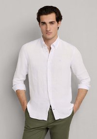 Muž oblečený v bílé košili na knoflíky s dlouhým rukávem, rukávy vyhrnuté, a olivově zelených kalhotách, s rukama v kapsách, před jednoduchým šedým pozadím.