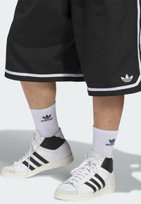 Mustat urheilushortsit, joissa on valkoinen reunus, yhdistettyinä valkoisiin korkeavartisiin lenkkikenkiin, joissa on mustia yksityiskohtia ja mustat Adidas-sukat.