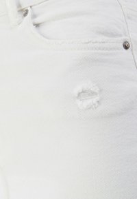 Närbild av vit denim med ett litet fransigt hål. Innehåller en ficka och en metallknapp som framhäver en avslappnad stil.