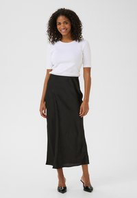 Jupe maxi en satin noir avec une texture lisse, taille haute et fente sur le côté ; assortie à un haut court à manches courtes en maille blanche et des talons noirs.