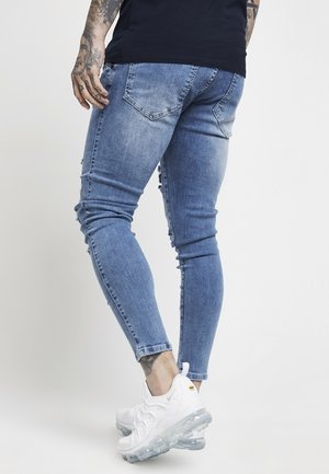 Man wearing faded skinny blue jeans and white sneakers, shown from the back against a plain light background.