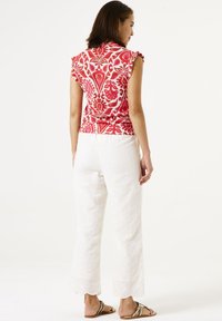 Top sin mangas de estampado rojo con hombros con volantes, combinado con pantalones de pierna ancha de ojo de perdiz blancos y sandalias planas, visto desde la parte trasera.