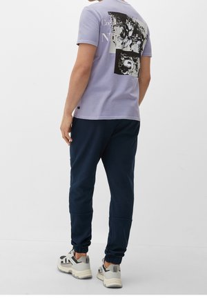 Mand iført en lyse lilla grafisk T-shirt, mørkeblå løbebukser og hvide og grå sneakers, stående mod en ensfarvet hvid baggrund.