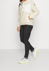 Sudadera de forro polar en color crema con un panel superior blanco, que cuenta con media cremallera y bolsillos frontales. Combinada con leggings negros y zapatillas plateadas con acentos en neón.