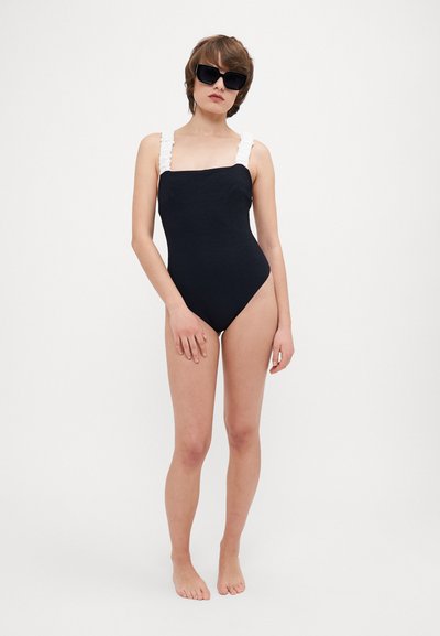 Black one-piece swimsuit with textured fabric, featuring white ruffled straps. Model wearing oversized black sunglasses, barefoot on a plain background.