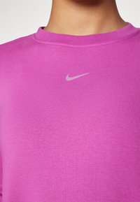 Camiseta Nike rosa con cuello redondo, hecha de tela suave. Presenta un pequeño logo de swoosh gris de Nike en el centro delantera. Textura suave.