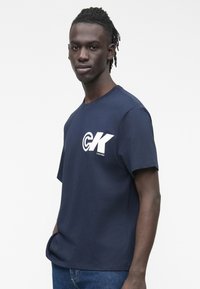 Man with short dreadlocks wearing a navy blue CK logo T-shirt and jeans, standing against a plain white background.